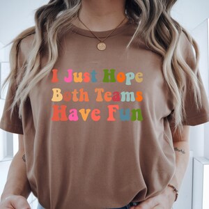 Peut inclure: T-shirt marron avec l'inscription "I Just Hope Both Teams Have Fun" dans des couleurs vives et un style r&eacute;tro. Le texte est en rouge, rose, orange, jaune, bleu et vert. Le t-shirt a des manches courtes et un col rond.