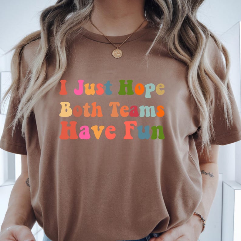 Peut inclure: T-shirt marron avec l'inscription "I Just Hope Both Teams Have Fun" dans des couleurs vives et un style r&eacute;tro. Le texte est en rouge, rose, orange, jaune, bleu et vert. Le t-shirt a des manches courtes et un col rond.