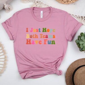 Peut inclure: T-shirt rose poudr&eacute; avec l'inscription &laquo; I Just Hope Both Teams Have Fun &raquo; dans une police color&eacute;e de style r&eacute;tro. Le t-shirt a des manches courtes et un col rond.