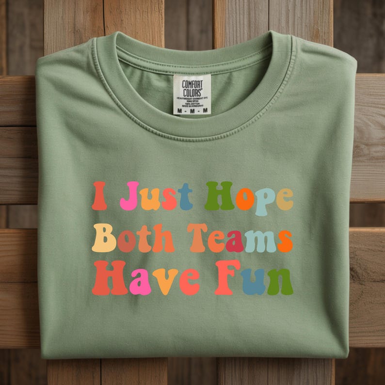 Peut inclure: T-shirt vert sauge avec l'inscription "I Just Hope Both Teams Have Fun" dans des lettres color&eacute;es de style r&eacute;tro. Le t-shirt a un col rond et une &eacute;tiquette Comfort Colors.