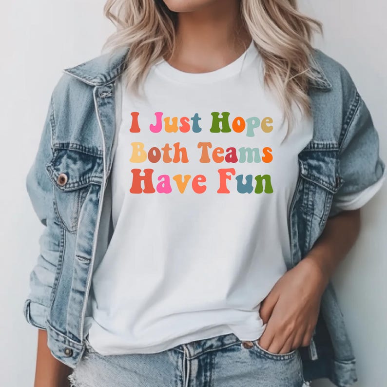 Peut inclure: T-shirt blanc avec le texte "I Just Hope Both Teams Have Fun" dans des lettres color&eacute;es de style r&eacute;tro. Le t-shirt est port&eacute; avec une veste en jean et un jean.