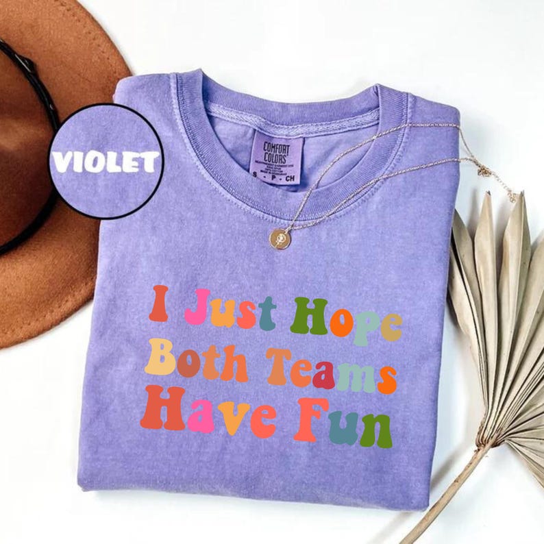 Peut inclure: Un t-shirt violet avec l'inscription "I Just Hope Both Teams Have Fun" en lettres color&eacute;es de style r&eacute;tro. Le t-shirt a un col rond et un collier dor&eacute; avec un pendentif. Un chapeau marron est partiellement visible en arri&egrave;re-plan.