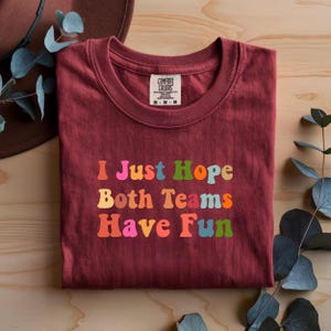 Peut inclure: Un t-shirt bordeaux avec l'inscription "I Just Hope Both Teams Have Fun" en lettres color&eacute;es de style r&eacute;tro. Le t-shirt a un col rond et une &eacute;tiquette "Comfort Colors".