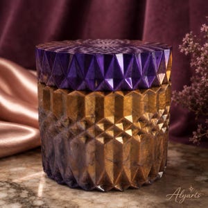 Puede incluir: Un recipiente cilíndrico con tapa, que presenta una textura superficial geométrica facetada. La tapa es de color morado intenso, mientras que la base transiciona de un dorado metálico a un morado oscuro. La superficie tiene un acabado marmolado.