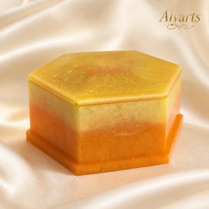 Puede incluir: Una caja hexagonal de resina con un esquema de color degradado que transita desde una parte superior de color amarillo pálido hasta una base de color naranja vibrante. La tapa tiene un acabado liso y brillante con partículas de purpurina.
