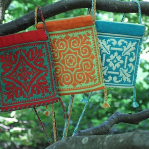 May include: Three rectangular crossbody bags with braided straps and tassels, featuring intricate geometric patterns. The first bag is red with teal details, the second is orange with light green details, and the third is teal with white details.