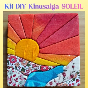 Op de afbeelding: Een vierkante Kinusaiga DIY-kit met een stoffencollage van een zon die opkomt boven een landschap. De zon is geel, omringd door rode en oranje stoffen stralen. Het onderste gedeelte toont bloemmotieven in roze, witte en blauwe stoffen stukjes.
