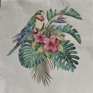 May include: A decorative textile featuring a parrot perched amidst tropical foliage. The parrot has green, blue, and yellow feathers, with a red patch on its chest. Pink hibiscus flowers and large green leaves complete the design.
