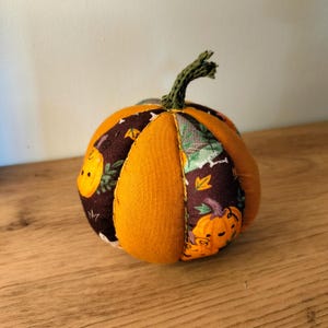 Peut inclure: Une citrouille en tissu rembourré composée de panneaux alternés en tissu orange uni et en tissu marron foncé imprimé de petites citrouilles orange et de feuilles vertes. La citrouille possède une tige crochetée vert foncé texturée.