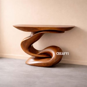 Puede incluir: Una consola de madera hecha a mano con un diseño único y fluido. La mesa presenta una tapa ovalada lisa sostenida por una base esculpida que se retuerce y curva. La madera tiene un acabado natural y cálido. El texto "CREAFYI" es visible.
