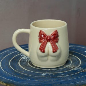 Puede incluir: Taza de cerámica color crema con un lazo rojo y dos formas redondeadas debajo. La taza tiene un asa y está sobre una superficie circular azul. El lazo es de un rojo vibrante.