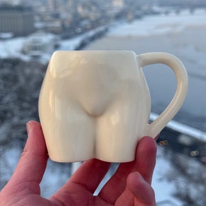 Puede incluir: Una taza de cerámica de color crema con forma de trasero humano. La taza tiene un asa redondeada y un acabado liso y brillante. El fondo muestra un paisaje urbano con un paisaje nevado.