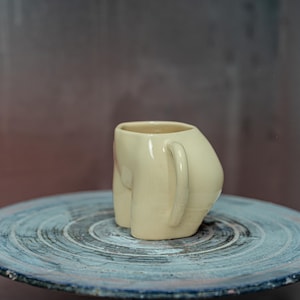 Pode incluir: Uma caneca de cerâmica creme com uma forma arredondada única, que lembra uma figura. A caneca tem uma alça curva e repousa sobre uma superfície circular azul-cinza. O fundo é um gradiente desfocado de castanho e cinza.