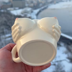 May include: Cream-colored ceramic mug shaped like a pair of breasts, with hands cupping the breasts. The nipples are painted pink. The mug has a handle and a round base.