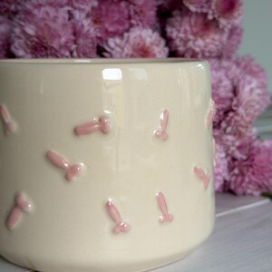 Puede incluir: Una taza de cerámica de color crema con un patrón repetitivo de pequeños adornos en relieve de color rosa claro con forma fálica en la superficie exterior. La taza tiene un acabado liso y brillante y una forma cilíndrica.