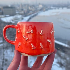 Könnte beinhalten: Ein leuchtend orangefarbener Keramikbecher mit glänzender Oberfläche, der mit einem wiederkehrenden Muster aus kleinen, erhabenen, cremefarbenen, phallusförmigen Tonverzierungen auf der Außenseite versehen ist. Der Becher hat einen geschwungenen Henkel.