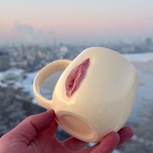 Puede incluir: Taza de cerámica color crema con un diseño de vulva rosa esculpida en el lateral. La taza tiene forma redondeada con asa. El fondo es un paisaje urbano borroso.