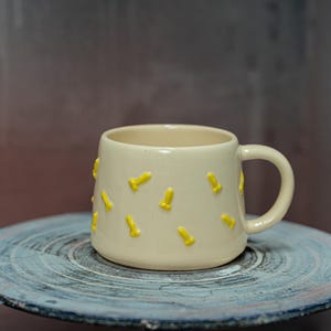 May include: Cream-colored ceramic mug with yellow, phallic-shaped embellishments. The mug has a rounded body, a curved handle, and sits on a round, blue-grey ceramic plate. The mug is a unique and quirky piece of drinkware.