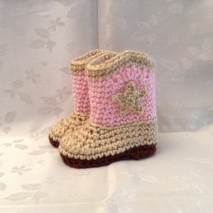 May include: A pair of handmade crochet baby cowboy boots. The boots are primarily beige and pink, with a brown sole. A beige star is stitched on the pink boot shaft. The boots are a charming, handcrafted item.