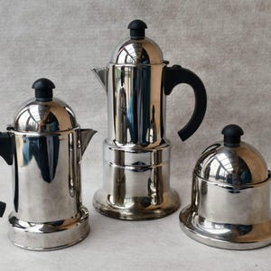 May include: Three stainless steel coffee makers with black handles and knobs. The coffee makers are different sizes and shapes, with a shiny, reflective surface. The background is a light, neutral color.