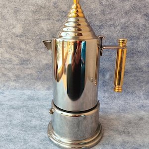 May include: A stainless steel stovetop espresso maker with a gold-colored handle and top. The coffee pot has a conical top and a round base. The handle and top are gold-colored, while the body is silver. The espresso maker is designed for brewing coffee.
