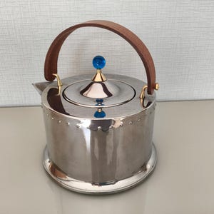 May include: A polished stainless steel teapot with a brown leather handle and a blue glass knob on the lid. The teapot has a round base and is decorated with small rivets around the body.