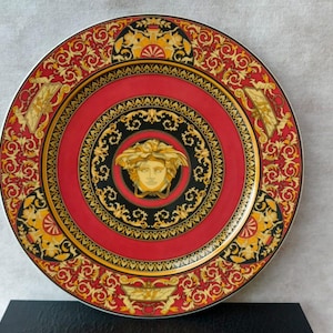 Puede incluir: Un plato decorativo con un centro rojo y detalles dorados y negros ornamentados. El plato presenta un emblema central de cabeza de Medusa y un borde estampado. El plato tiene forma redonda y se muestra sobre una superficie negra.