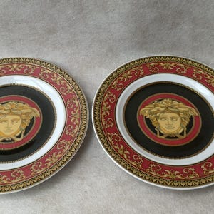 May include: Two decorative plates with a black center featuring a gold-colored face design. The plates have a red and gold border with a white inner ring. The plates are round and have a gold rim.