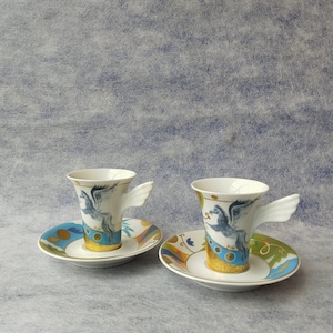 May include: Two white ceramic espresso cups with wing-shaped handles, each resting on a matching saucer. The cups and saucers feature a blue and gold design with a winged horse motif. The saucers have a colorful, patterned border.