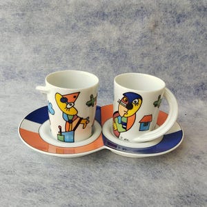 May include: A set of two white ceramic coffee cups with abstract, colorful illustrations. Each cup sits on a matching saucer, connected to form a single unit. The saucers feature a blue, white, and orange striped design.