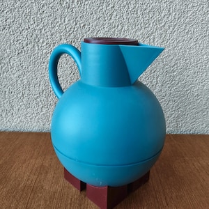 May include: A teal-coloured, spherical jug with a handle and a spout. The jug has a dark brown lid and sits on a dark red base. The design is modern and minimalist, suitable for serving beverages.