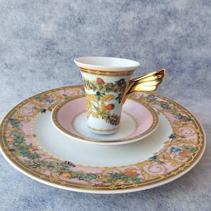 May include: A white porcelain teacup with a gold wing-shaped handle, set on a matching saucer and a larger plate. The cup and plates feature a floral and fruit design with gold trim and a pink border. The set is designed for serving tea.