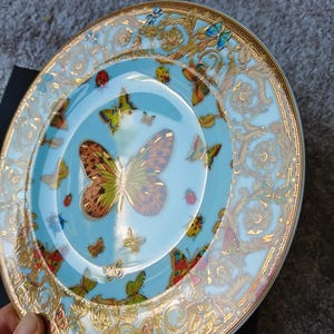 May include: A decorative plate with a light blue centre and a gold rim. The plate features a butterfly design with various colours and patterns. The rim has a gold ornate design.