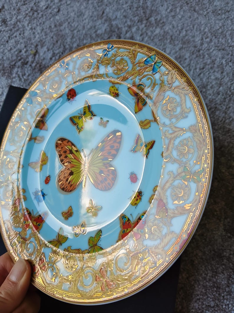 May include: A decorative plate with a light blue centre and a gold rim. The plate features a butterfly design with various colours and patterns. The rim has a gold ornate design.