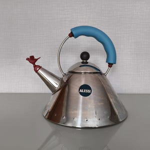 May include: A stainless steel Alessi kettle with a conical body, a light blue curved handle, and a small red bird-shaped whistle on the spout. The kettle features a black circular logo with the text ALESSI on the front and a black knob on the lid.