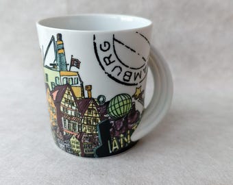 May include: White ceramic mug with a colourful illustration of a cityscape, including buildings, a crane, and a globe. The mug has a curved handle and a black stamp that says "HAMBURG".