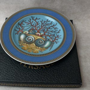 May include: A decorative plate with a blue and gold rim, featuring a central design of seashells and coral in shades of blue, gold, and red. The plate rests on a black box with a gold border.