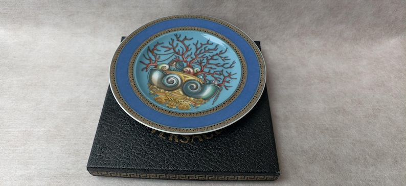 May include: A decorative plate with a blue and gold rim, featuring a central design of seashells and coral in shades of blue, gold, and red. The plate rests on a black box with a gold border.