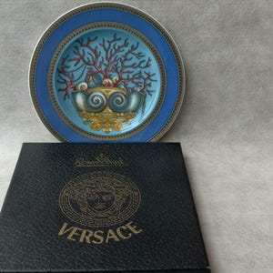 May include: A decorative plate with a blue rim and a central design featuring a coral-like motif in shades of red, teal, and gold. The plate rests above a black box with the gold Versace logo.