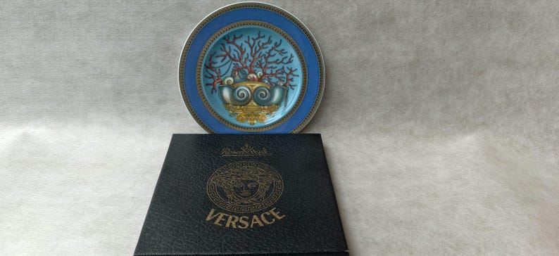 May include: A decorative plate with a blue rim and a central design featuring a coral-like motif in shades of red, teal, and gold. The plate rests above a black box with the gold Versace logo.