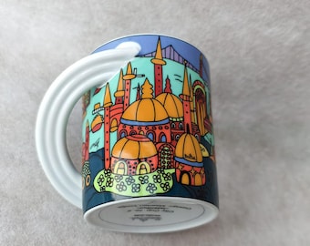 May include: A ceramic mug with a white handle and a colourful illustration of a cityscape. The design features buildings with orange domes, red towers, and a blue sky. The mug has a white handle.