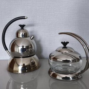 May include: Two stainless steel teapots with black handles and black knob details. One teapot is solid stainless steel, and the other has a glass body. Both have a modern, minimalist design.