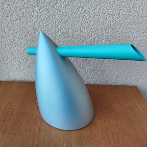 May include: A modern, abstract teapot in a light blue color with a turquoise spout. The teapot has a unique, angular design and sits on a wooden surface. The spout extends horizontally from the body.