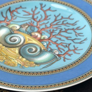 May include: A decorative plate with a light blue border and a central design featuring seashells, coral, and gold accents. The plate has a detailed pattern around the edge and a turquoise center.