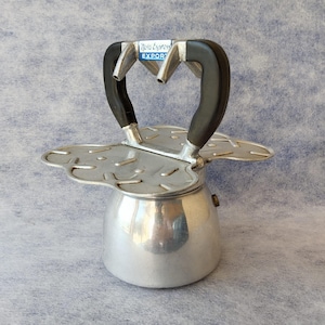 Puede incluir: Una cafetera italiana vintage plateada con asas negras. La cafetera tiene un diseño único con un asa en forma de corazón y el texto "Nova Express Export" en una etiqueta azul.