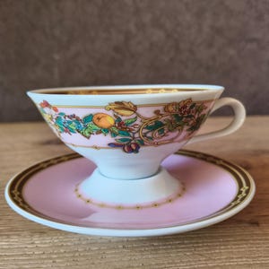 May include: A pink teacup and saucer set with a gold rim and floral design. The teacup has a white base and a handle. The saucer is pink with a gold border. The set is on a wooden surface.