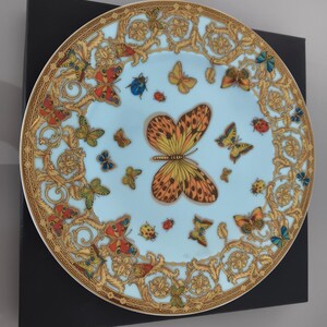 May include: A decorative plate with a light blue background, adorned with a butterfly and insect design. The plate has a gold ornate border and is presented on a black surface. The butterflies are in various colours, including orange, yellow, and blue.