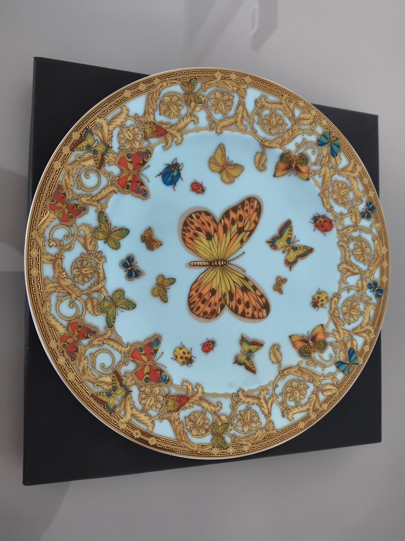 May include: A decorative plate with a light blue background, adorned with a butterfly and insect design. The plate has a gold ornate border and is presented on a black surface. The butterflies are in various colours, including orange, yellow, and blue.