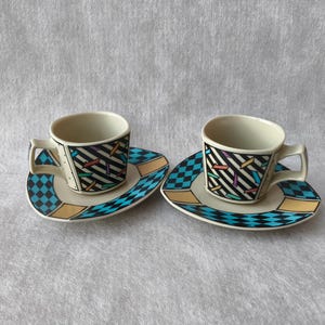 Könnte beinhalten: Zwei Espressotassen und Untertassen aus Keramik. Die Tassen und Untertassen haben eine cremefarbene Basis mit geometrischen Mustern in Schwarz, Blau, Gelb und verschiedenen Farben. Die Untertassen sind herzförmig.