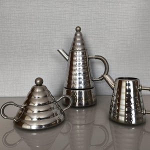 May include: A set of three silver-toned coffee and tea serving pieces. The set includes a coffee maker, a creamer, and a sugar bowl. Each piece has a textured, hammered finish and a coiled handle. The coffee maker is approximately 10 inches tall.